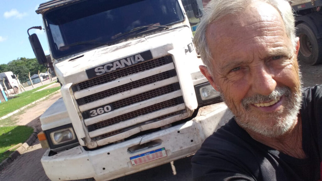 Caminhoneiro sorridente tira selfie em frente a seu caminhão Scania branco, estacionado em um pátio ao ar livre.