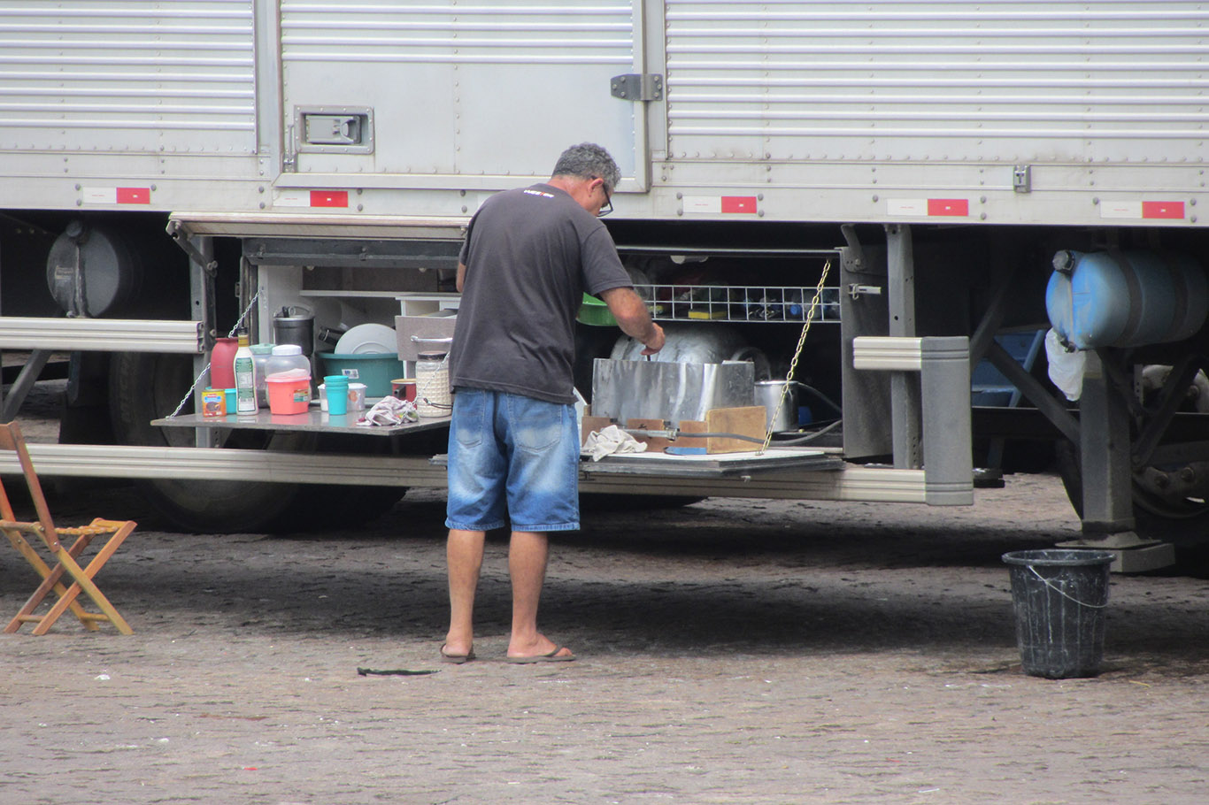 Homem em frente a caminhão preparando comida em cozinha improvisada montada na traseira do veículo, com utensílios, balde e cadeira dobrável ao redor.