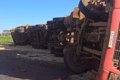 Caminhão tombado sobre a lateral em uma estrada, com a carroceria exposta e rodas visíveis.