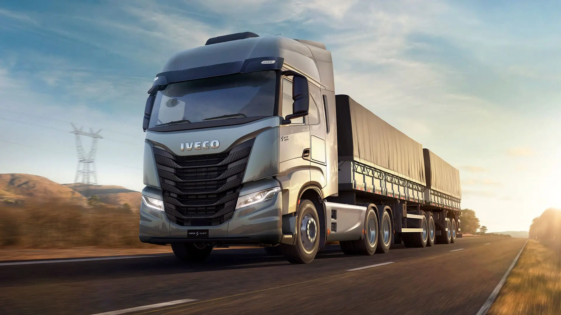 Caminhão Iveco S-Way prata puxando carreta com lonas, em movimento em uma rodovia ao entardecer.