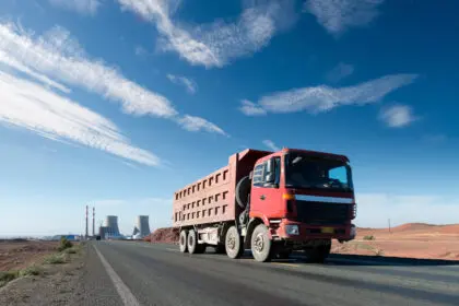Caminhão basculante vermelho trafegando por uma estrada asfaltada em uma paisagem árida, com céu azul e nuvens finas; ao fundo, instalações industriais com torres e chaminés.