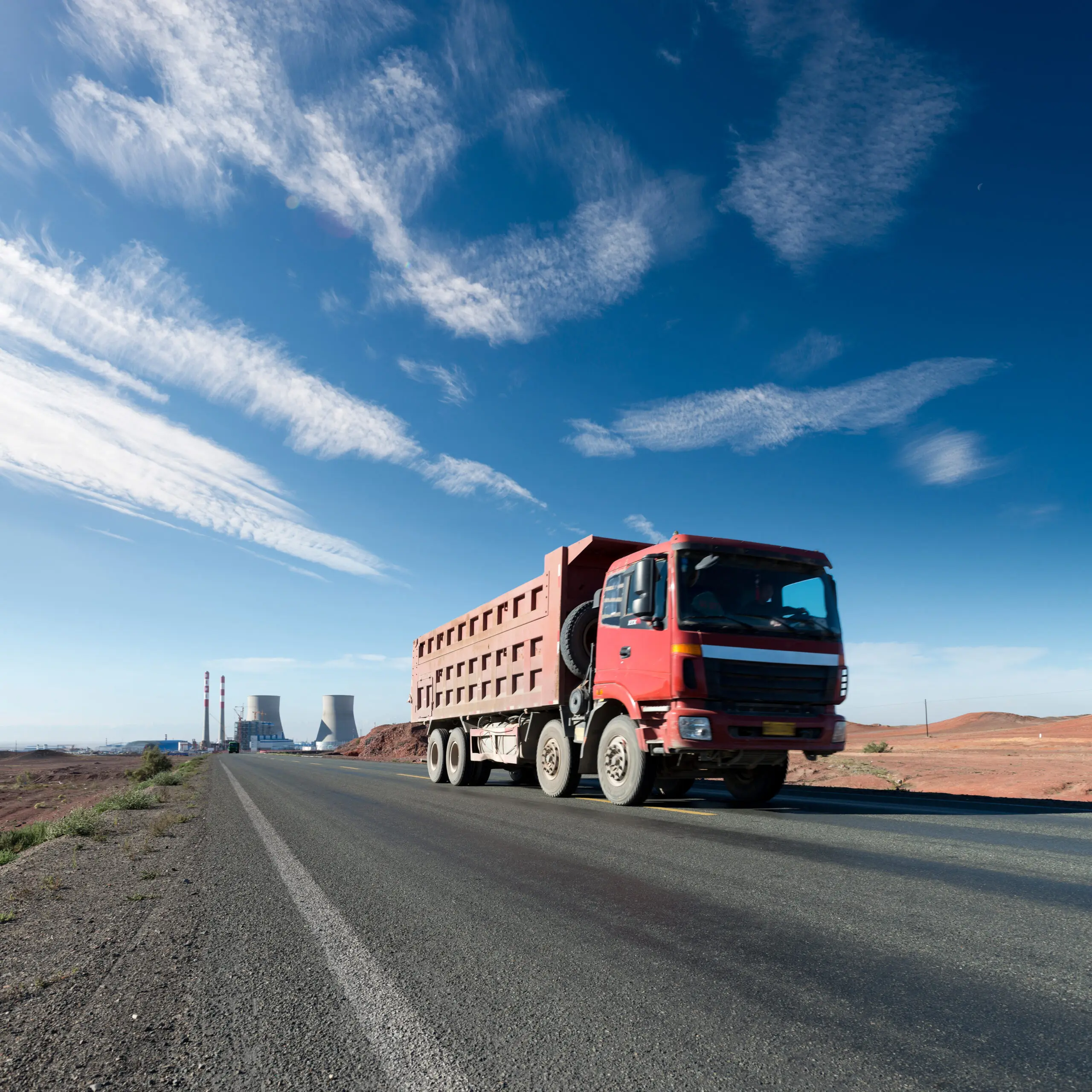 Caminhão basculante vermelho trafegando por uma estrada asfaltada em uma paisagem árida, com céu azul e nuvens finas; ao fundo, instalações industriais com torres e chaminés.