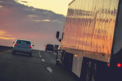 Caminhão e carros trafegam em rodovia ao entardecer, com o pôr do sol refletido na lateral do veículo de carga.
