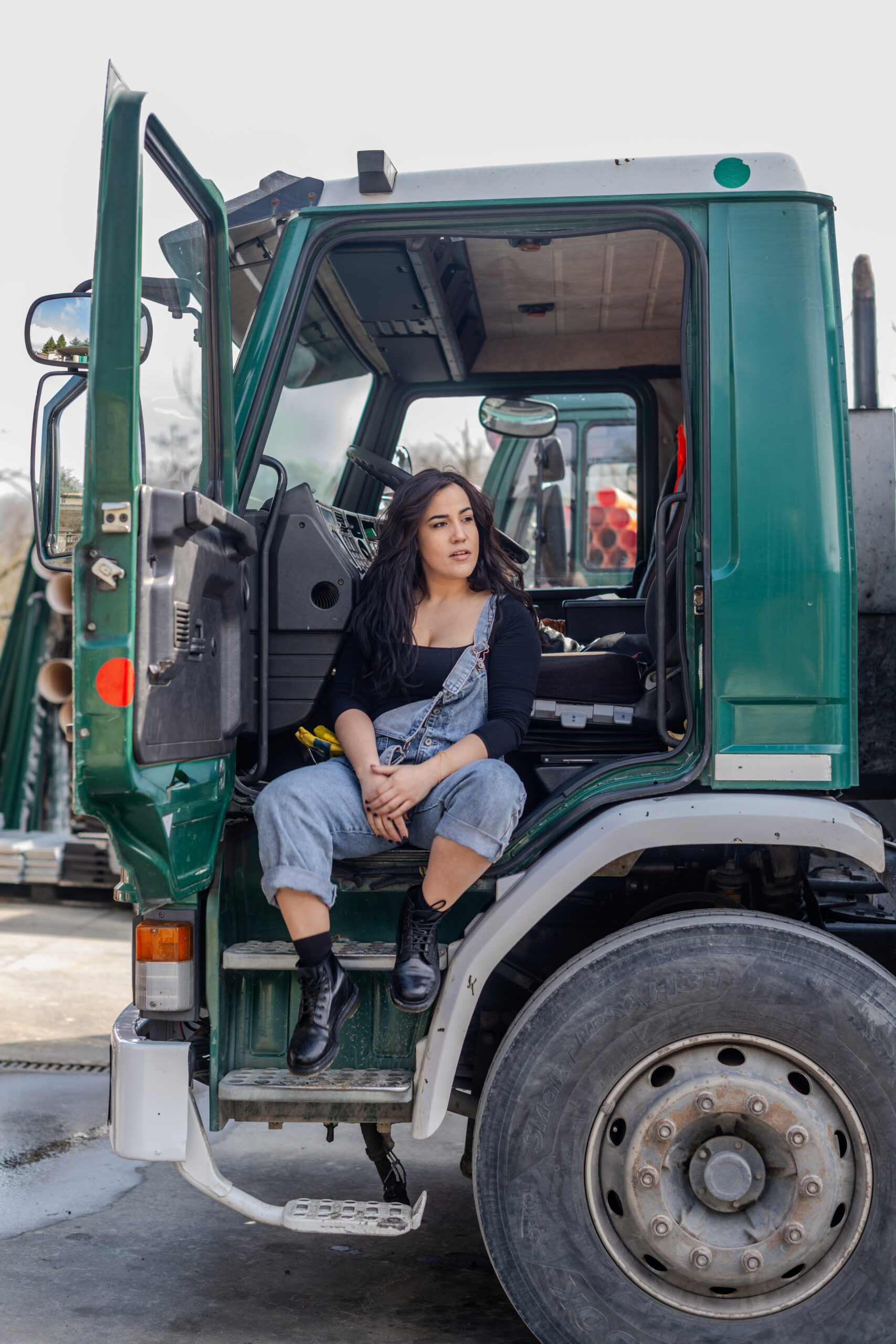 Hispanic young model poses in black shirt and blue overalls lean up set a big green cargo truck