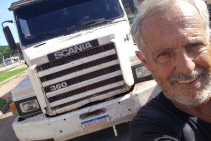 Caminhoneiro sorridente tira selfie em frente a seu caminhão Scania branco, estacionado em um pátio ao ar livre.