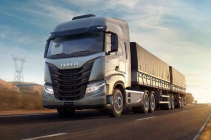 Caminhão Iveco S-Way prata puxando carreta com lonas, em movimento em uma rodovia ao entardecer.