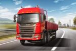Caminhão JAC vermelho 6x2 trafegando em rodovia com paisagem de montanhas e prédios ao fundo.
