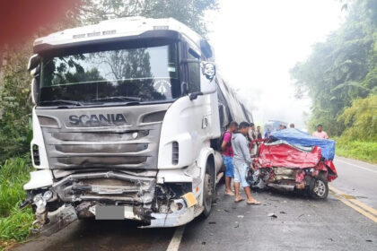 Caminhão Scania colidido com carro vermelho fortemente destruído na frente, em rodovia de pista simples, com pessoas observando o acidente.