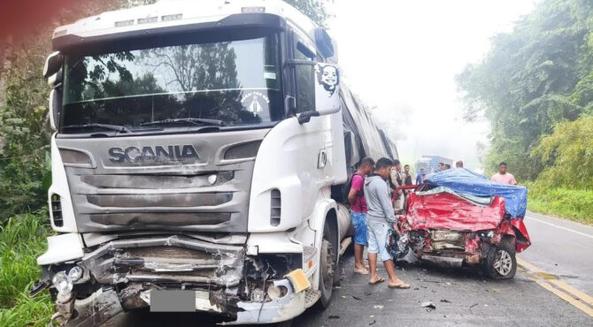 Caminhão Scania colidido com carro vermelho fortemente destruído na frente, em rodovia de pista simples, com pessoas observando o acidente.