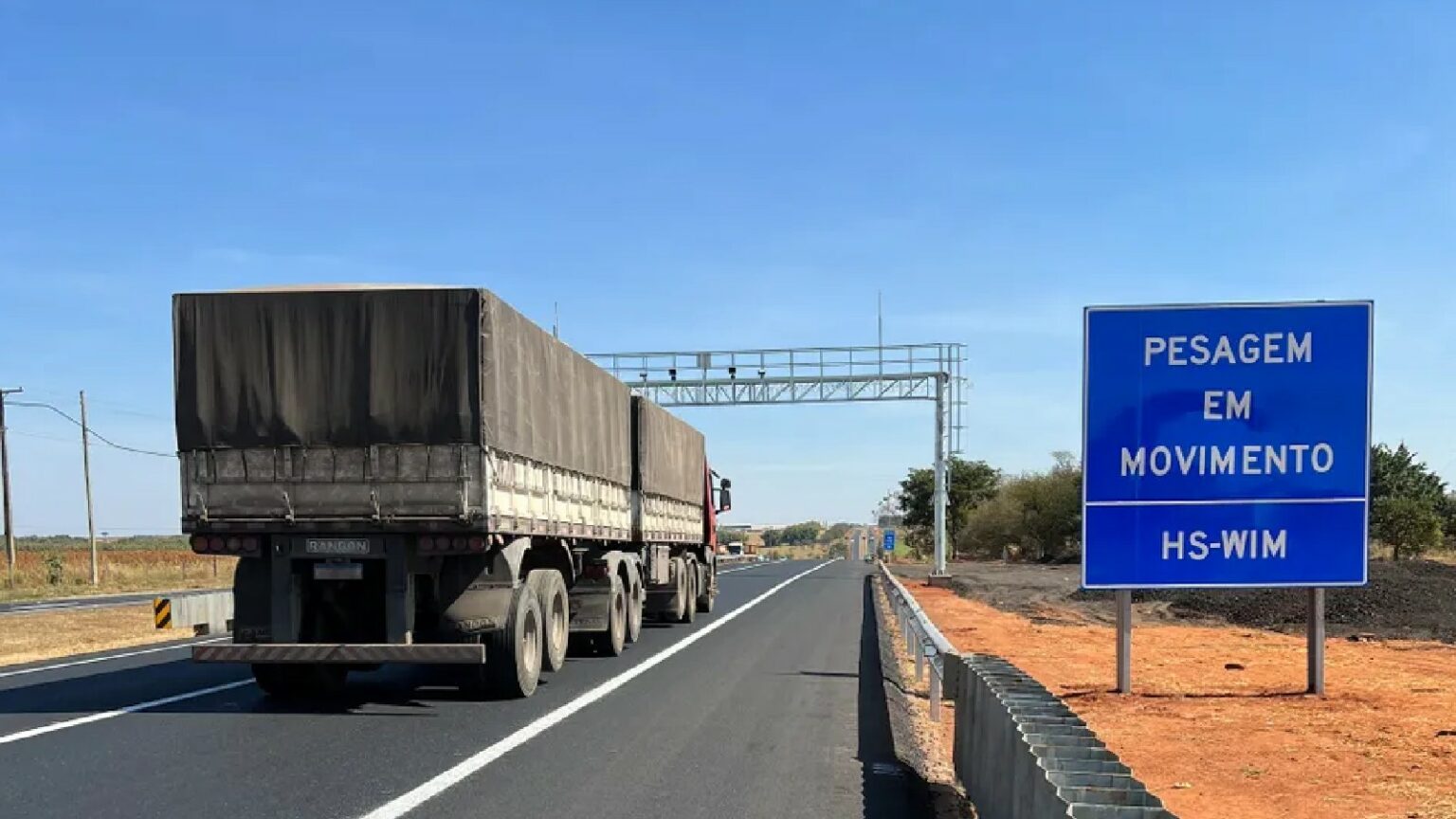 Caminhão em rodovia passando por sistema de pesagem em movimento indicado por placa azul.