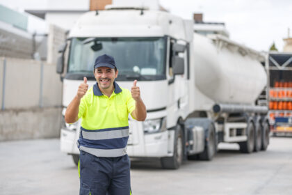 Caminhoneiro sorridente com uniforme refletivo em frente a caminhão tanque branco, mostrando sinal positivo com os polegares.