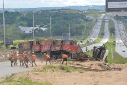 Caminhão tombado às margens de uma rodovia, com vários trabalhadores usando coletes de segurança realizando atendimento no local.