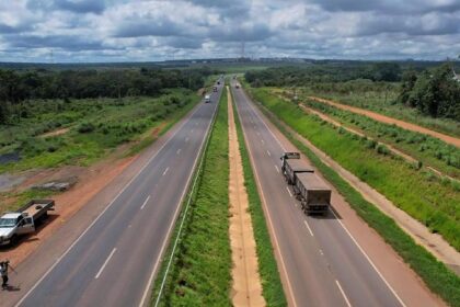 Rodovia duplicada corta área rural, com veículos trafegando nos dois sentidos entre faixas de vegetação.