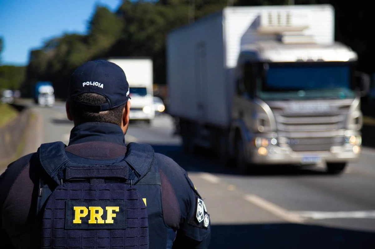 Agente da Polícia Rodoviária Federal, visto de costas, observa caminhões trafegando em rodovia.