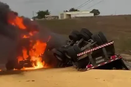 Caminhão tombado em uma estrada, com chamas intensas e fumaça ao fundo.