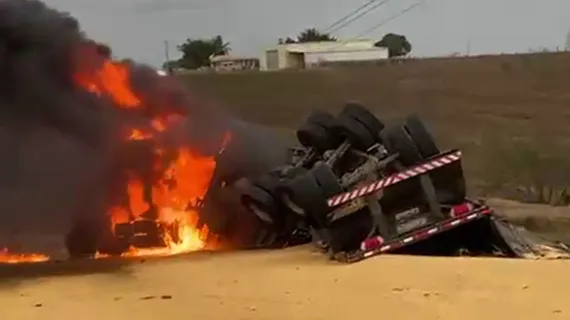 Caminhão tombado em uma estrada, com chamas intensas e fumaça ao fundo.