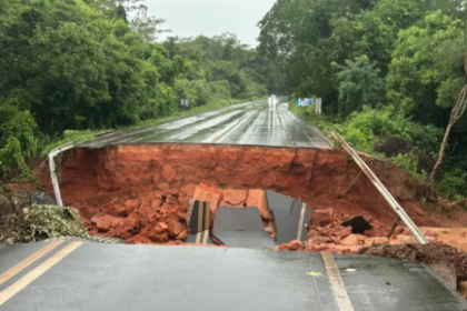 Trecho de rodovia em área de mata interrompido por um grande deslizamento de terra, com parte do asfalto desabado e a pista completamente rompida sob chuva.