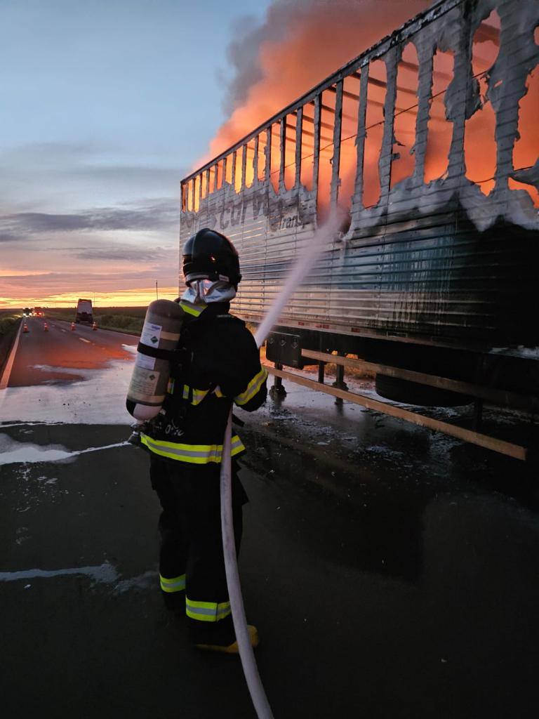 Bombeiro combate incêndio em carreta na beira de rodovia ao entardecer, com chamas e fumaça saindo da lateral do veículo.