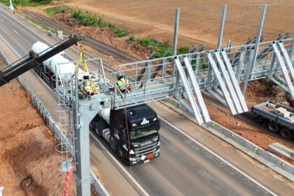 Trabalhadores com equipamentos de segurança instalam uma estrutura metálica para free flow sobre uma rodovia, usando um guindaste, enquanto um caminhão passa sob a obra.