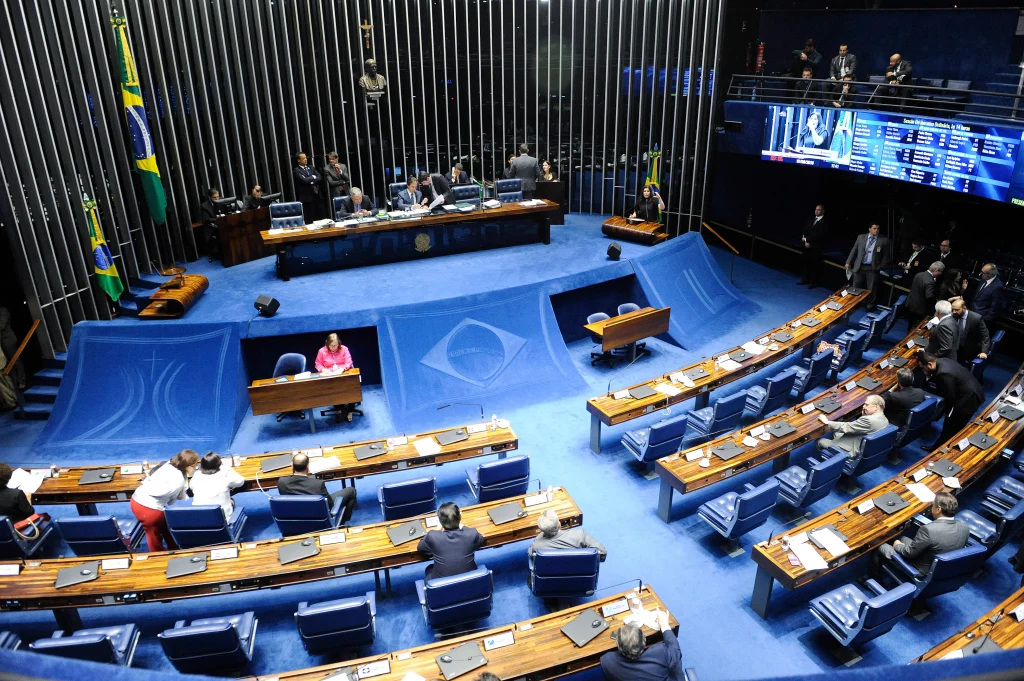 Plenário do Senado com mesas e cadeiras dispostas em semicírculo, com poucos parlamentares presentes e espaço central vazio.