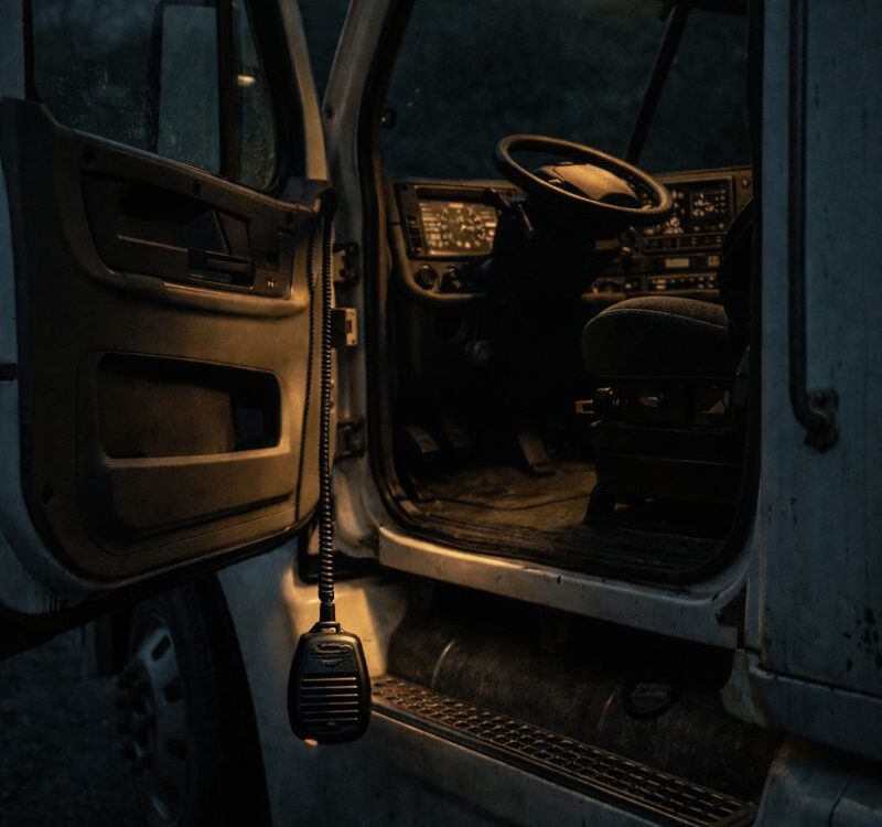 Interior da cabine de um caminhão à noite, com a porta aberta, volante visível, painel iluminado e um rádio comunicador pendurado ao lado do banco do motorista.