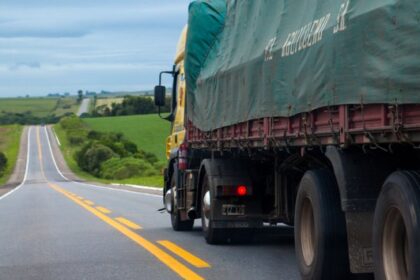 Caminhão de carga trafega por rodovia de pista simples em área rural, com campos verdes ao redor e céu nublado.