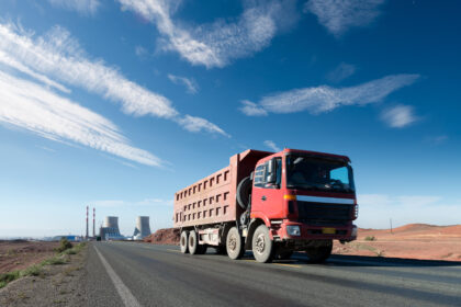 Caminhão basculante vermelho trafegando por uma estrada asfaltada em uma paisagem árida, com céu azul e nuvens finas; ao fundo, instalações industriais com torres e chaminés.