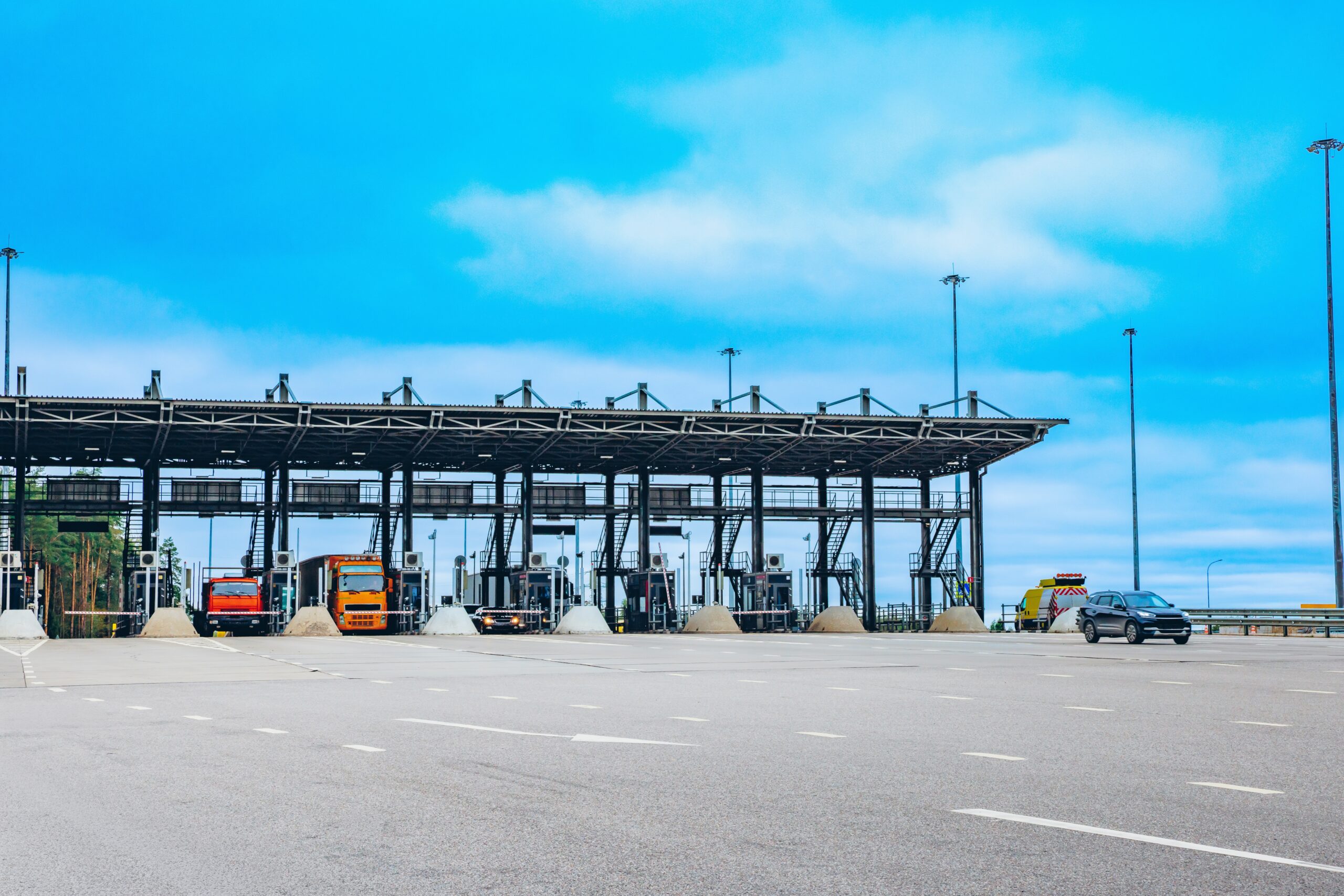 Praça de pedágio em rodovia com várias cabines, caminhões e carros parados ou em circulação, sob céu azul.