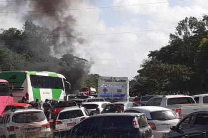 Trânsito congestionado em rodovia com vários veículos parados e fumaça escura ao fundo, próxima a caminhões e carros.