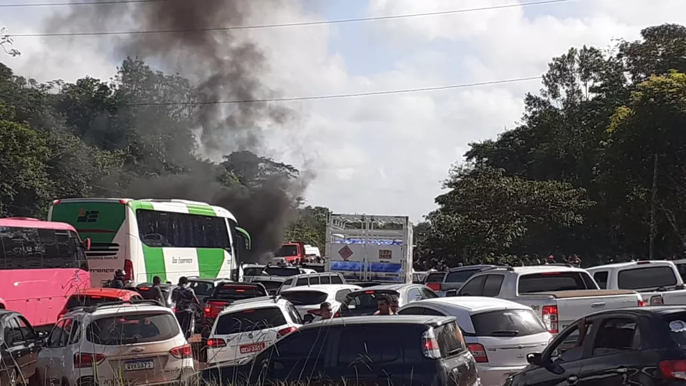 Trânsito congestionado em rodovia com vários veículos parados e fumaça escura ao fundo, próxima a caminhões e carros.