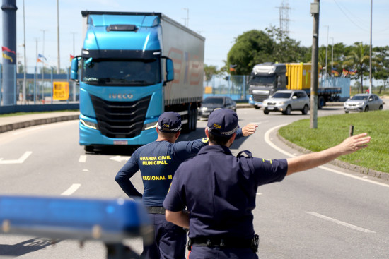 Agente de trânsito sinaliza parada de caminhão azul em via urbana durante fiscalização.