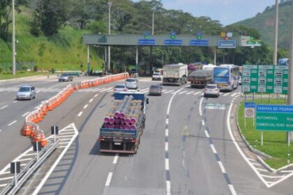 Caminhões e carros trafegam por rodovia com várias faixas, separados por cones laranja, em trecho urbano.