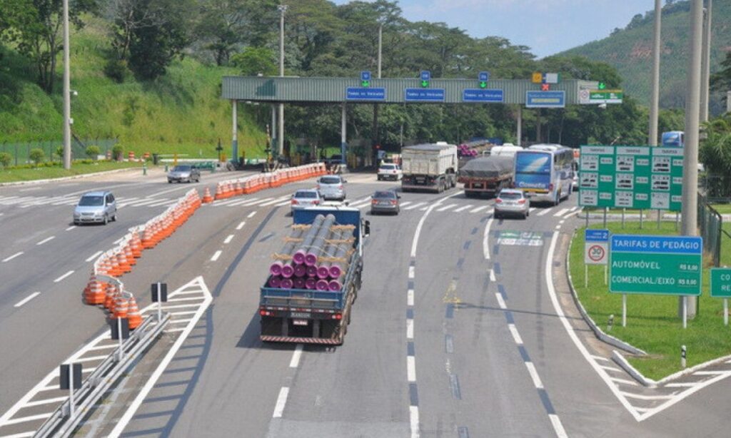 Caminhões e carros trafegam por rodovia com várias faixas, separados por cones laranja, em trecho urbano.