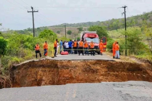 Grupo de trabalhadores com coletes laranja ao lado de um caminhão em área de obra, diante de uma cratera no solo.