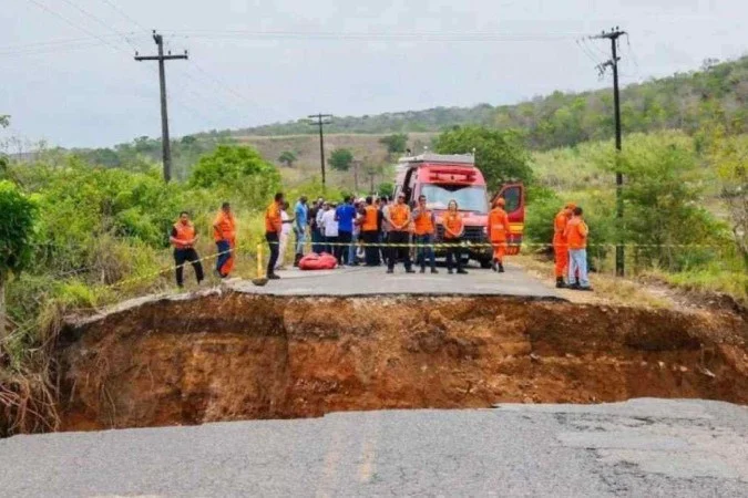 Grupo de trabalhadores com coletes laranja ao lado de um caminhão em área de obra, diante de uma cratera no solo.