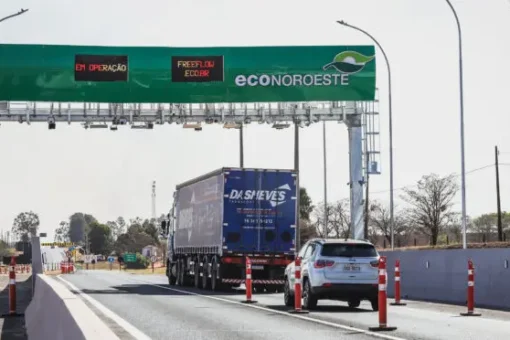Caminhão azul com contêiner passando por pórtico de pedágio free flow em rodovia.