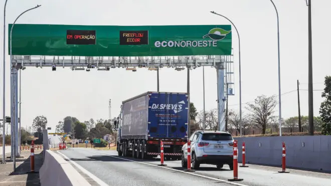Caminhão azul com contêiner passando por pórtico de pedágio free flow em rodovia.