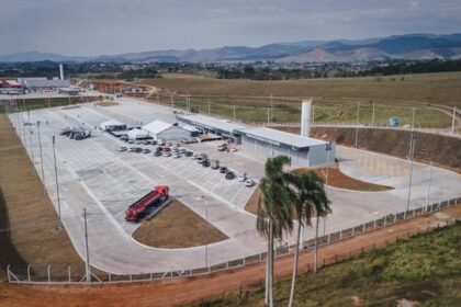 Vista aérea de um grande posto rodoviário com amplo pátio pavimentado, vagas de estacionamento, caminhão-tanque, carros estacionados e prédio principal, cercado por área rural e montanhas ao fundo.