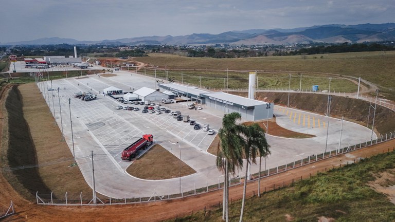 Vista aérea de um grande posto rodoviário com amplo pátio pavimentado, vagas de estacionamento, caminhão-tanque, carros estacionados e prédio principal, cercado por área rural e montanhas ao fundo.