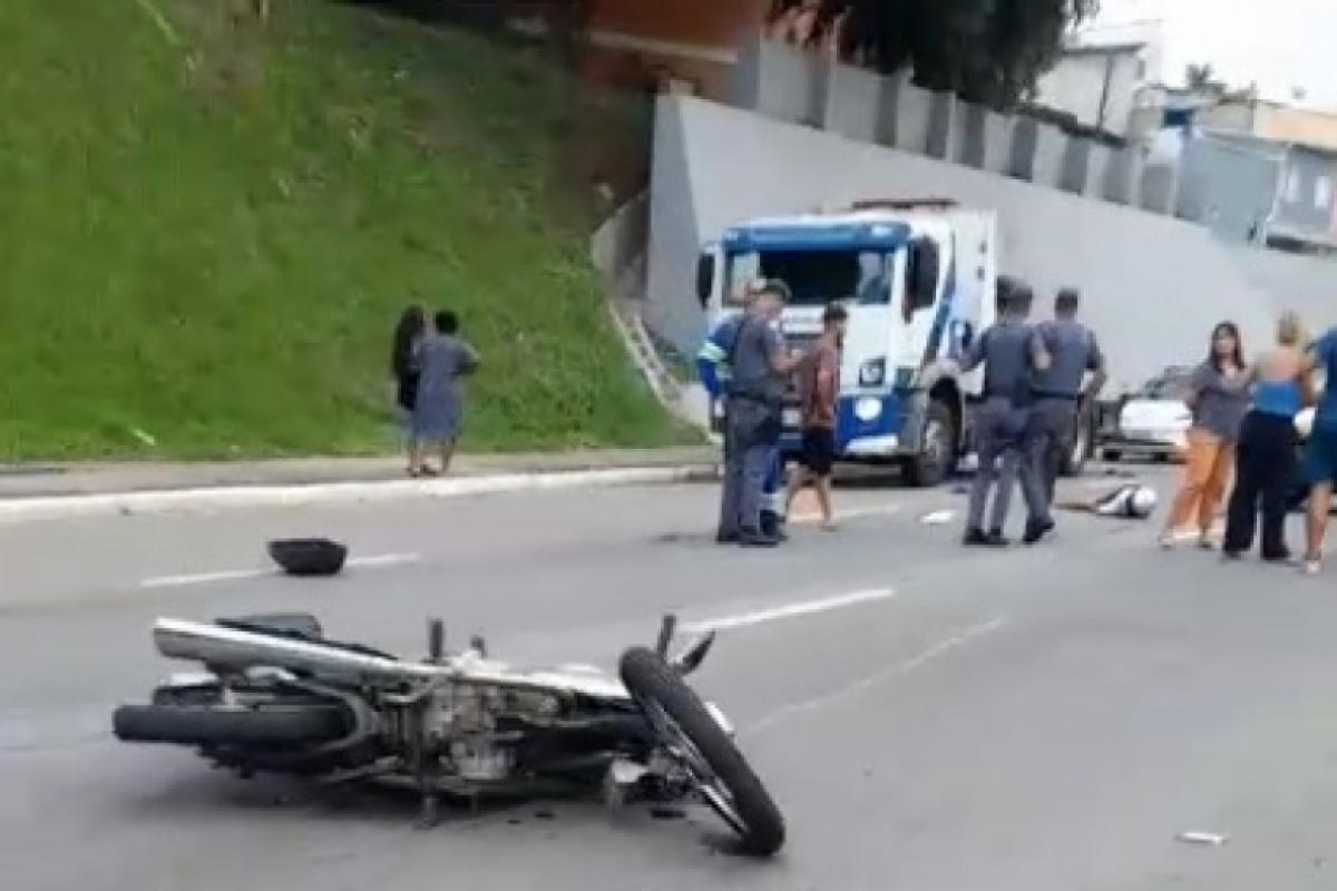 Motocicleta caída no asfalto em via urbana, enquanto policiais e equipes de resgate atendem ocorrência de acidente próximo a uma ambulância.