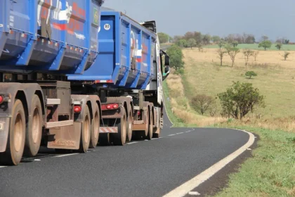 Caminhão azul de grande porte trafega por uma estrada asfaltada e sinuosa em área rural, com vegetação seca ao redor.