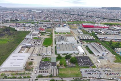 Vista aérea de um complexo industrial com vários galpões, áreas verdes e vias internas, cercado por bairro urbano ao fundo.