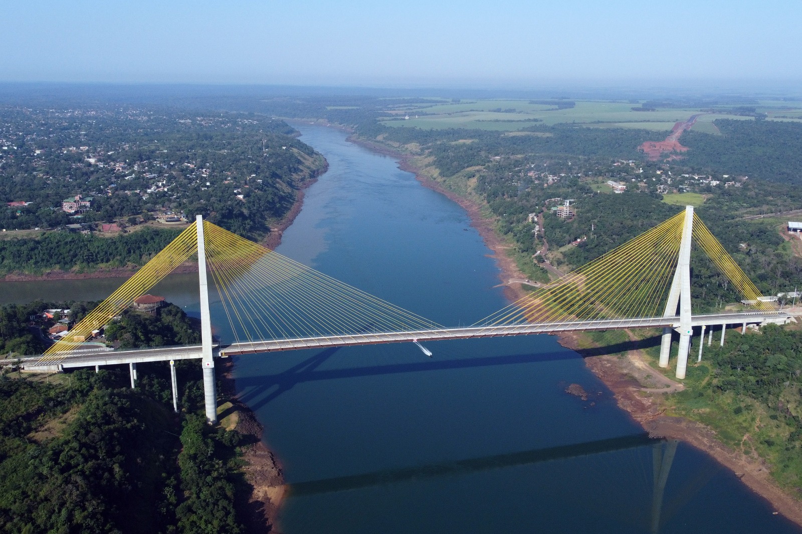 Vista aérea de uma ponte estaiada atravessando um rio largo, com cabos amarelos e vegetação nas margens.