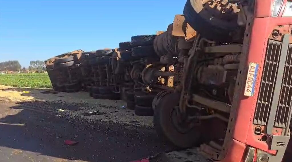 Caminhão tombado sobre a lateral em uma estrada, com a carroceria exposta e rodas visíveis.