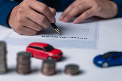 Mão segurando uma caneta ao lado de carrinhos em miniatura e pilhas de moedas sobre uma mesa.