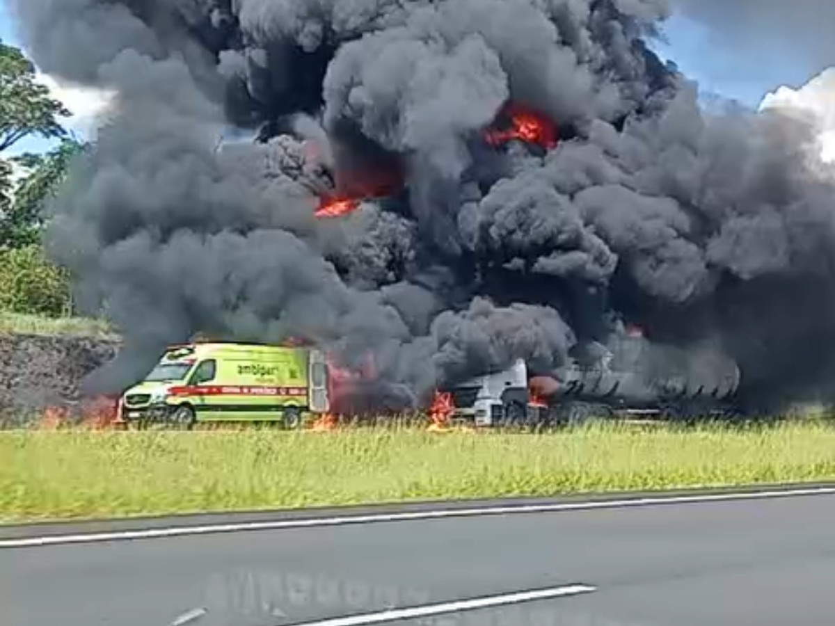 Caminhão em chamas às margens de uma rodovia, com intensa fumaça preta se espalhando pelo céu.