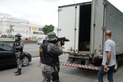 Agente de segurança armado aponta fuzil em direção à parte traseira aberta de um caminhão estacionado na rua.