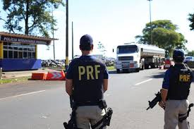 Agente da Polícia Rodoviária Federal, visto de costas, usando colete com a sigla PRF, em uma rodovia com caminhões ao fundo.