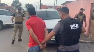 Homem com camisa vermelha é conduzido por um agente de segurança em via pública.