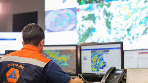 Profissional de costas, usando uniforme laranja, observa mapas meteorológicos exibidos em duas telas de computador.